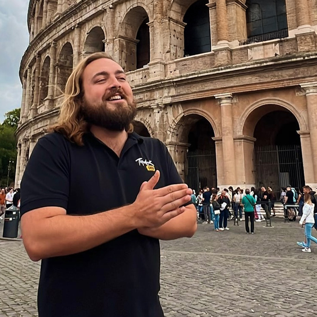 Man with beard clasps his hands together while smiling in front of the Colosseum in Rome.