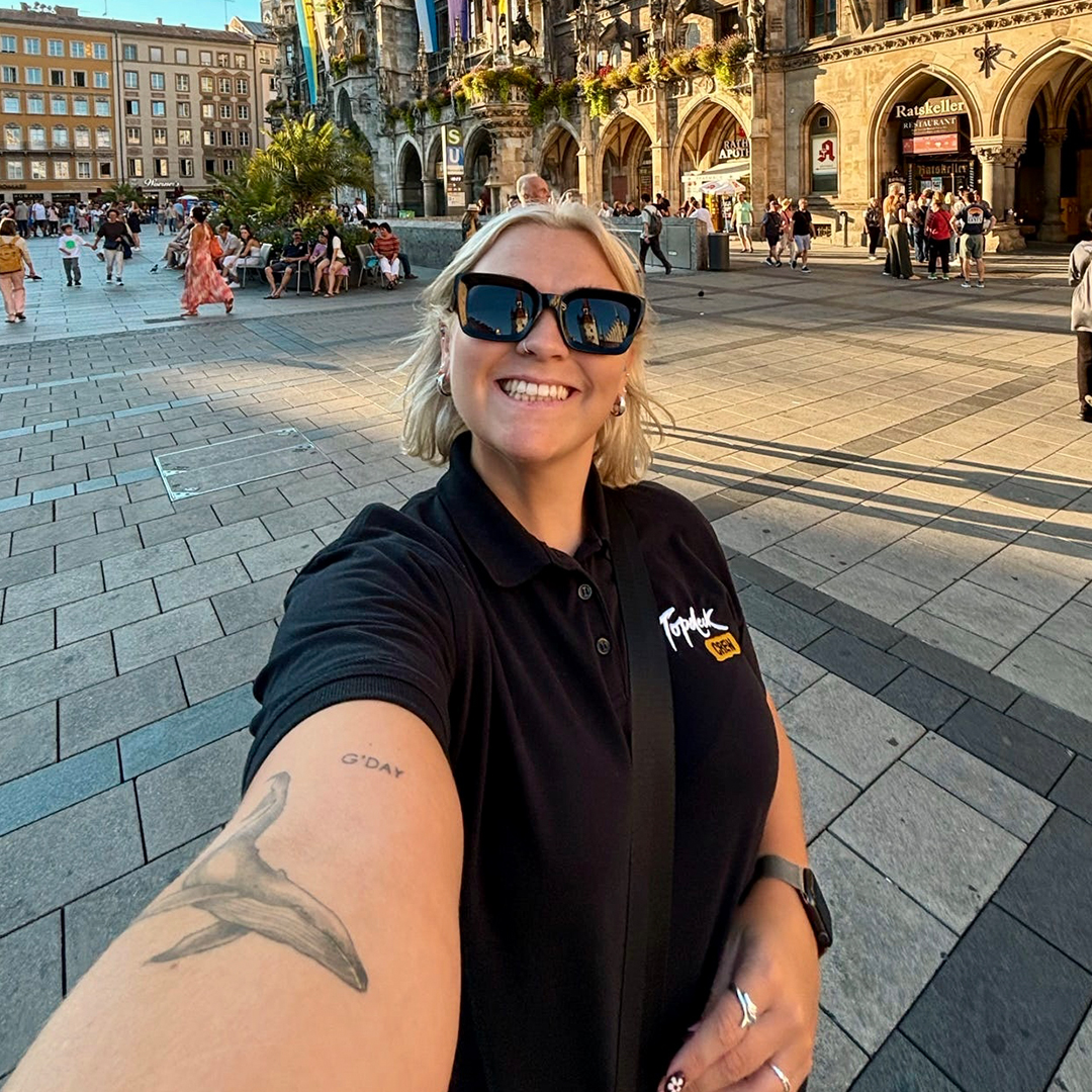 Woman with blonde hair and sunglasses on holds her arm out while taking a selfie in front of a European building.
