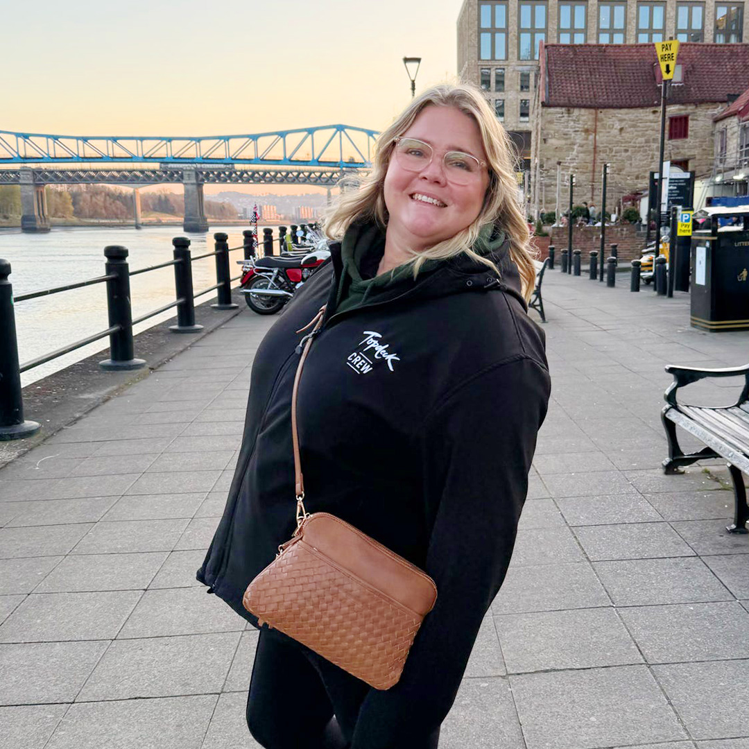Woman with blonde hair and glasses on stands in front of a bridge with a brown cross body bag on her side.