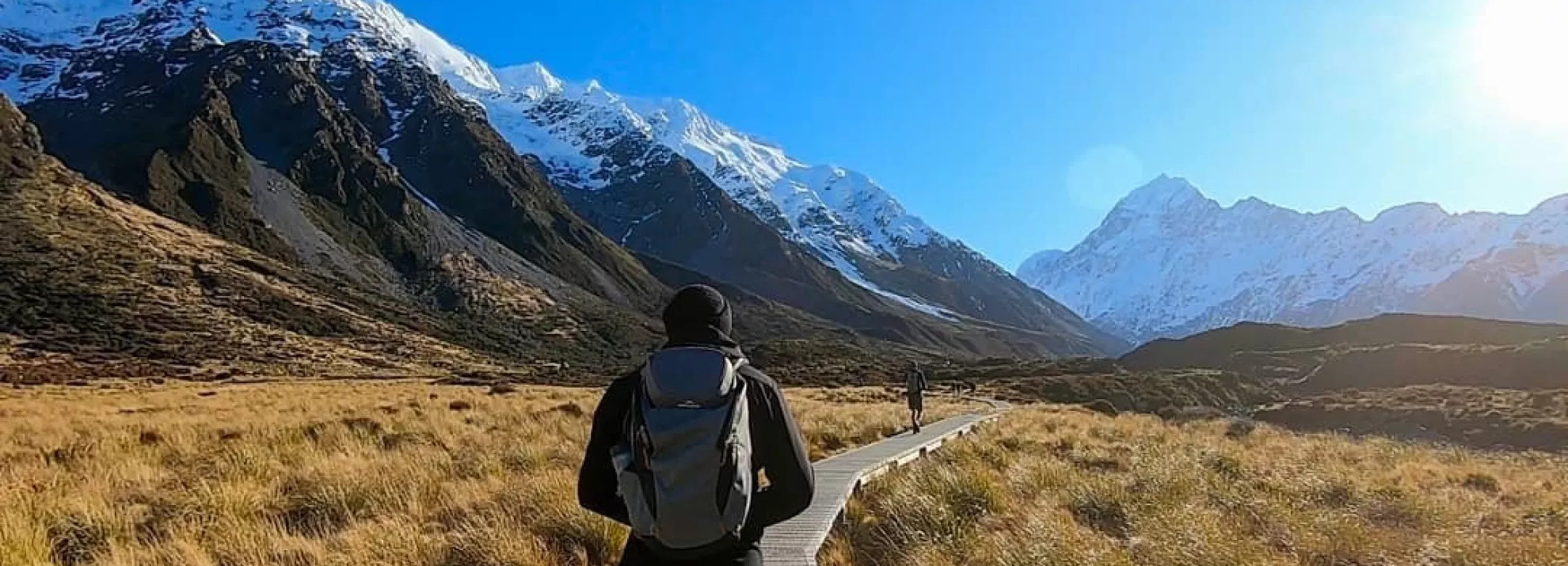 Man walking down a walkway with mountains and a bright blue sky in the background