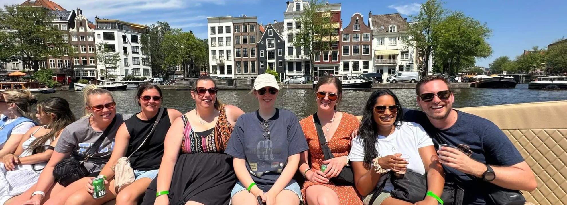 Group of six women and one man all wearing sunglasses and smiling at the camera with a background view of the river