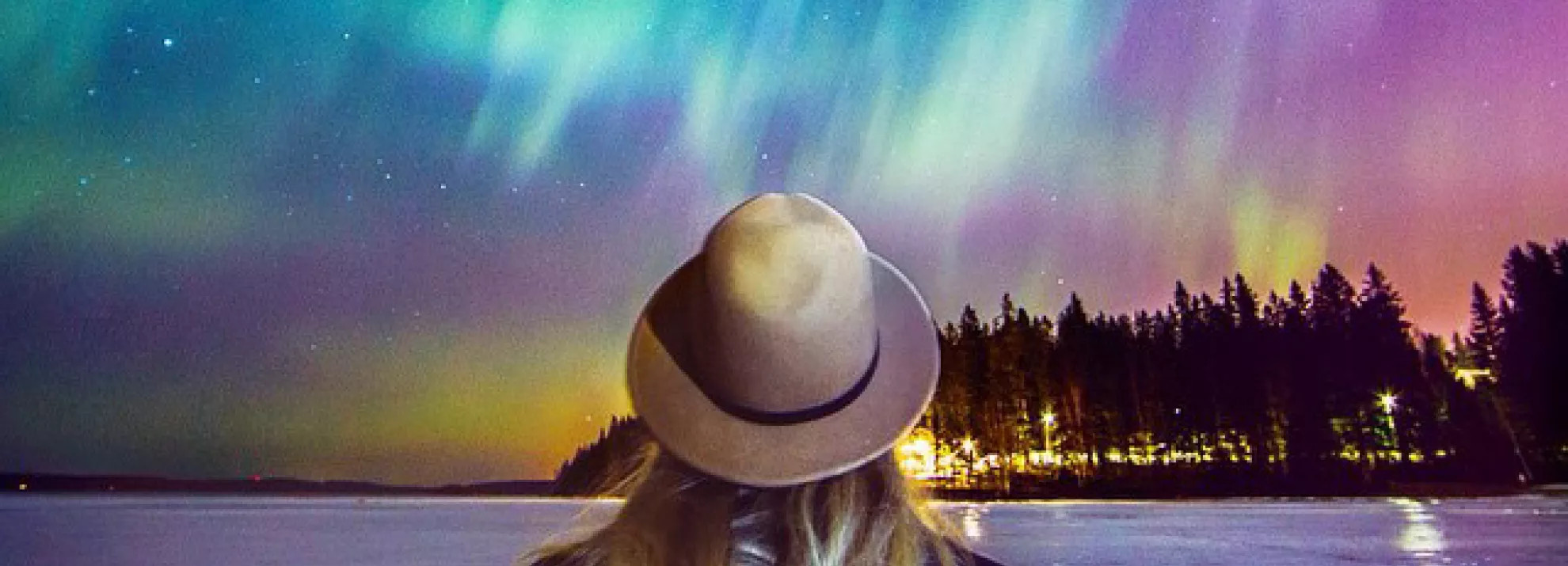 Woman stands with her back to the camera wearing a hat. The background is of the northern lights