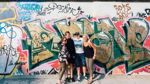 Four people standing together smiling in front of the graffitied Berlin Wall.