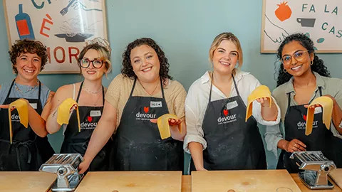 Five people holding sheets of pasta with two pasta machines and wooden boards in front of them.