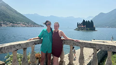 Two people standing together against a stone fence with a lake behind them.