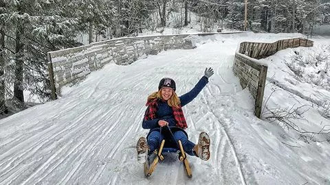 Person wearing winter clothing sits on a taboggan on a snow covered track.