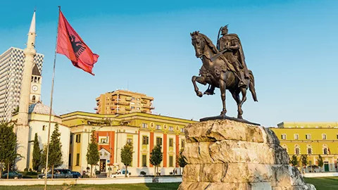 Statue of man riding a horse and Albanian flag flying near a large building.