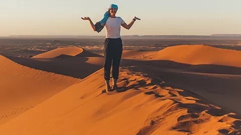 Person stands on the ridge of a large sand dune with their hands in the air.
