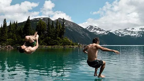 Two people jump into a Canadian lake with snow capped mountains in the distance.