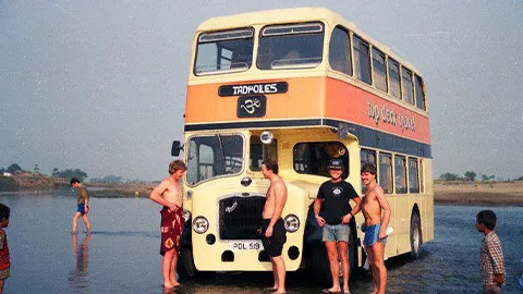 People standing in front of an orange double decker original Topdeck bus on a beach.