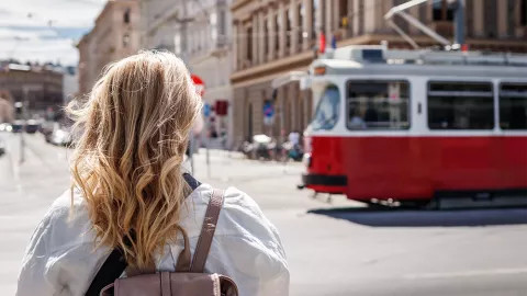 Person with blonde hair standing on the street as a red and white tram passes by.