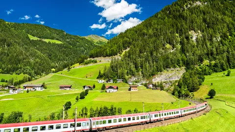 Train runs along the Brenner Railway through grassy hills with houses in the distance with red rooves.