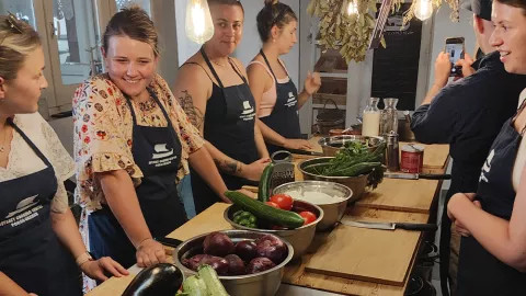 Group of people stand around a table wearing aprons for a Greek cooking class. There are vegetables in bowls in the center of the table.