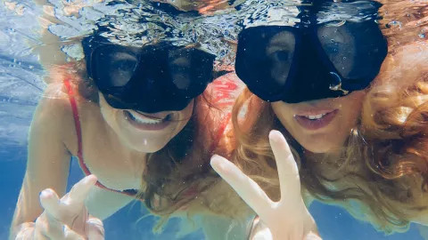 Two girls smiling underwater while wearing a snorkel mask and making peace signs with their hands.