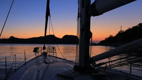 View of sailing boat’s bow at dusk as the sun sets over the hills in the distance.