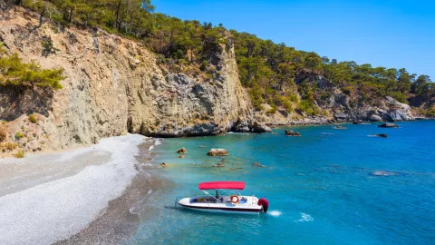 A small boat with red roof sits against the stoney shore in turquoise water on Ölüdeniz Beach.