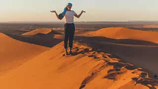 Woman standing smiling on sand dune ridge with her arms out and palms facing the sky.