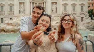 Three people smile while holding out coins in front of the Trevi Fountain.