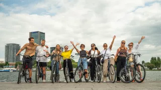 Group of people smiling with their hands in the air. They are sitting on bikes near the river in Amsterdam.
