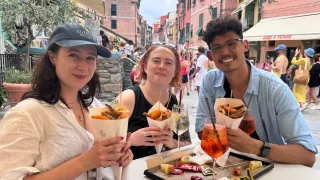 Three people smiling at a table in a Venice street. They hold fried street food and have spritz drinks in front of them.