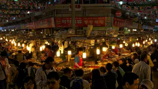 Corner of indoor Gwangjang Market, dimly lit with laterns. Woks of traditional Korean food are laid out with customers ordering their meals.