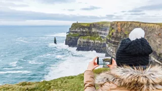 Person wearing a beanie takes a photo on their phone of the white water along the coastline.