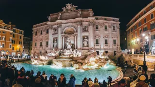 The famous Trevi Fountain of Rome lit up at night with a crowd of people surrounding it.