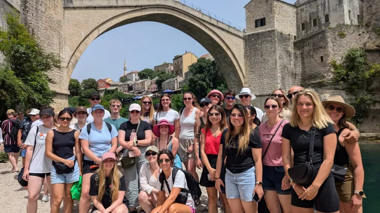 A group of topdeckers standing in front of an old bridge 