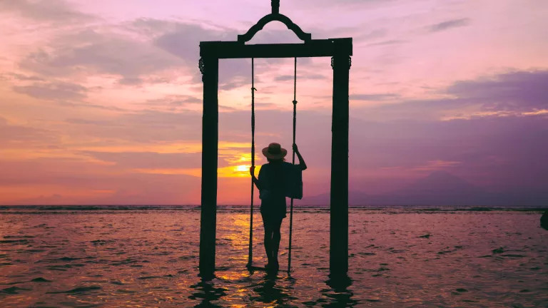 A woman standing on a swing in the ocean, while the sunsets