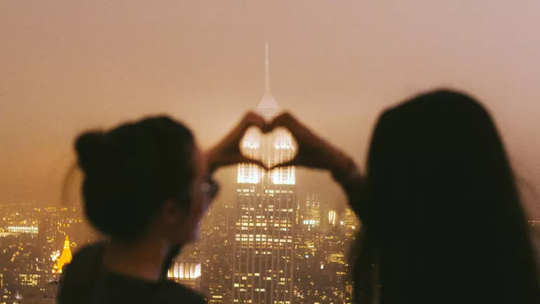 Two women holding their hands together to form a heart with the Empire State Building in the background
