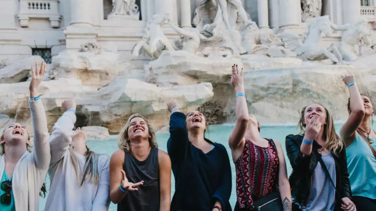 Happy group of people throwing a coin behind them into the Trevi Fountain