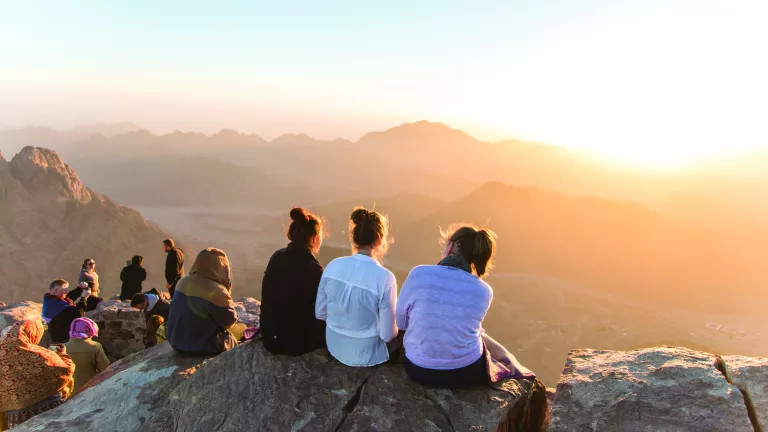 Three women sitting on a cliff looking out at mountains