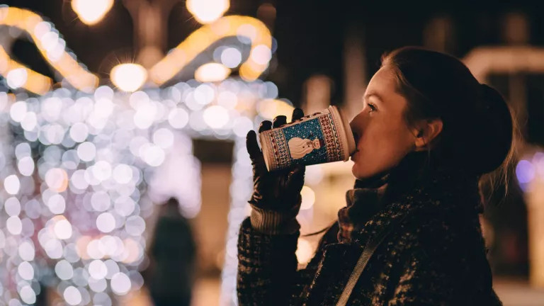 woman drinking a hot drink at a christmas market. There are blurry lights in the background