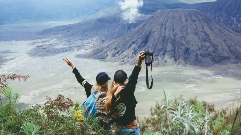 Two women in an embrace, holding their arms up together. In the background, they look over a volcano