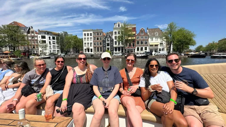 Group of six women and one man all wearing sunglasses and smiling at the camera with a background view of the river