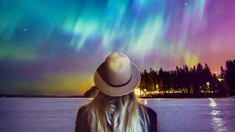 Woman stands with her back to the camera wearing a hat. The background is of the northern lights