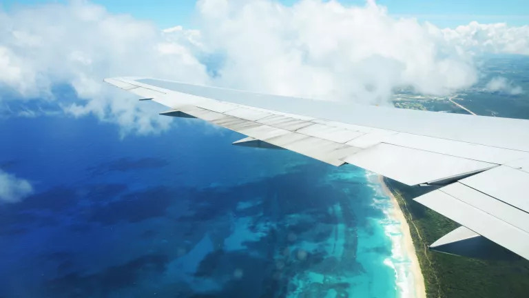 View outside a plane window. You can see the wing of the plane on the right and a beautiful blue ocean on the left
