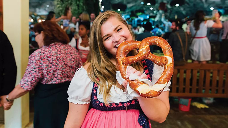 Person wearing a dirndls holding a large pretzel at an Oktoberfest celebration.