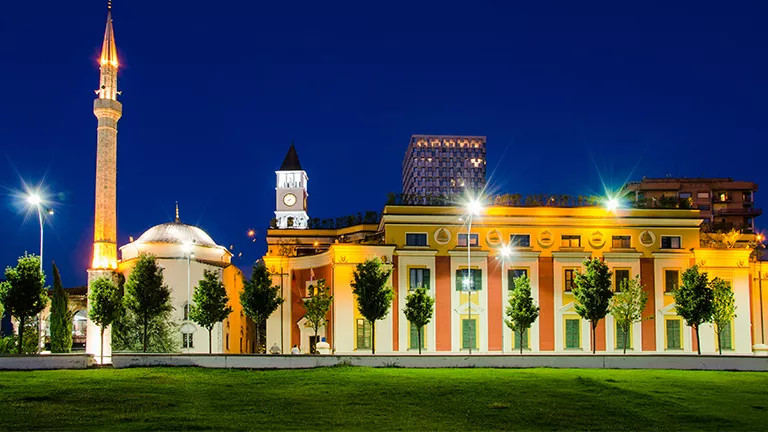 Building with bright outdoor lights at night with large spire and clock tower nearby.