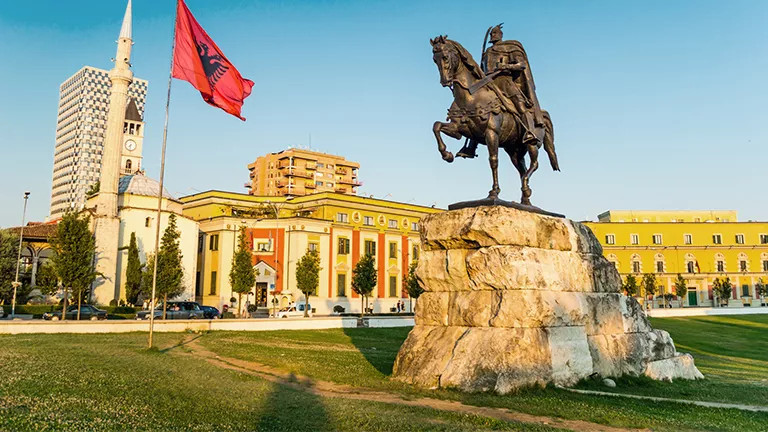 Statue of a main on a horse with the Albanian flag flying nearby in front of buildings.