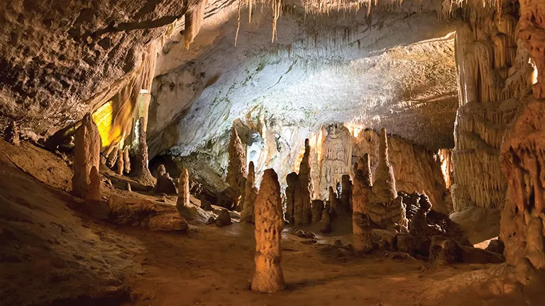 Cave with large stalactites and stalagmites lit up by light.