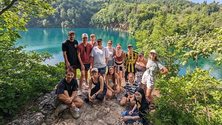 Group poses together on a large rock in a national park, surrounded by lush forest and blue water.