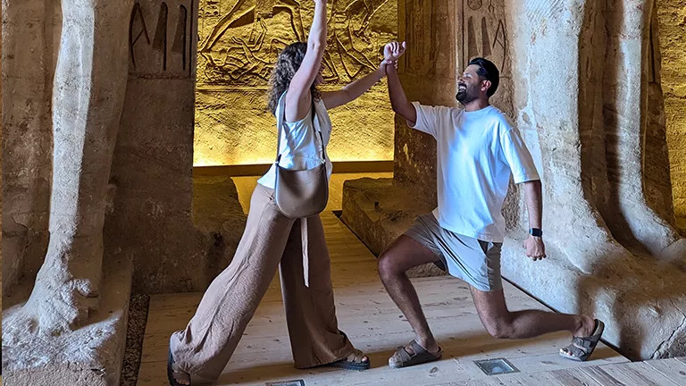 Two people strike a playful pose inside an Egyptian temple. There are hieroglyphic like designs on the wall behind them.