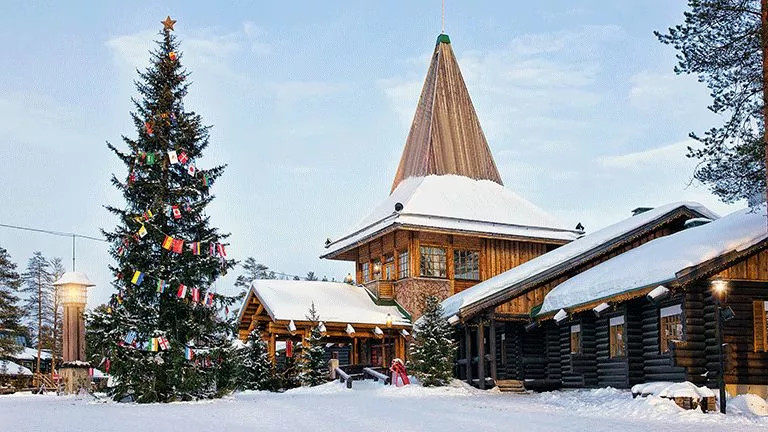 Building covered in snow with pointed roof and decorated Christmas tree in the early evening.