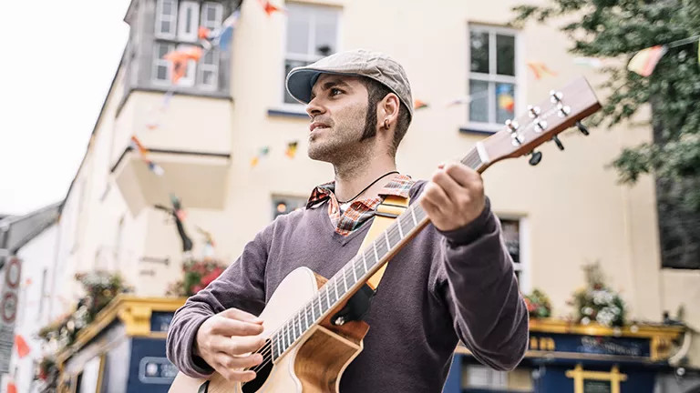 Person wearing a collared shirt and cap playing an acoustic guitar outside a pub.