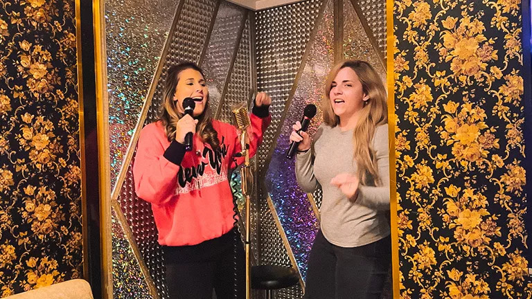 Two people holding microphones sing karaoke against a glittery wall.