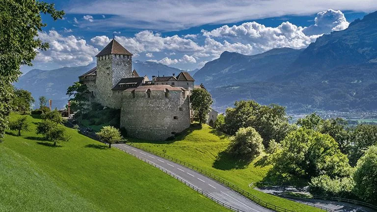 Stone walls of Vaduz Castle with a rolling green hill in front and mountains behind.