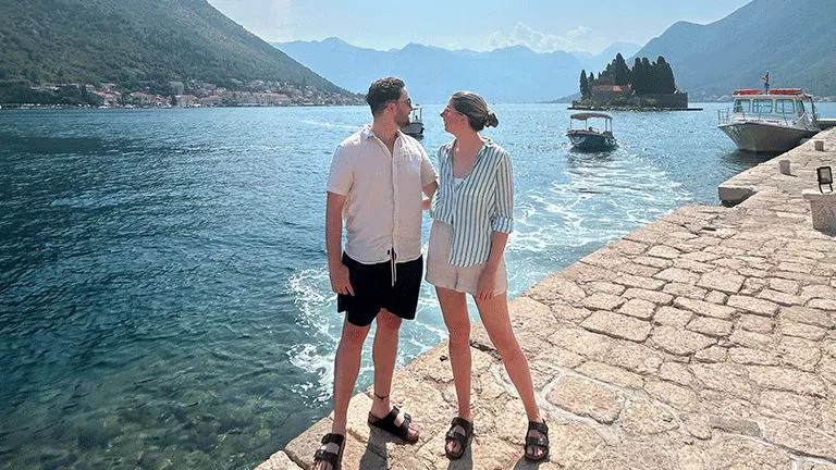 Two people standing on a stone paved walkway next to the water.