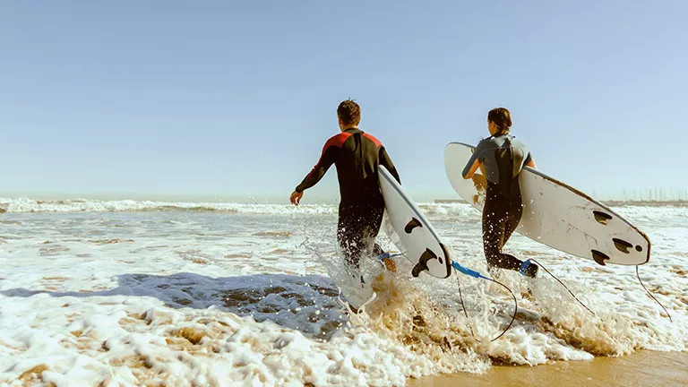 Two people stepping into waves on the beach while carrying surf boards.
