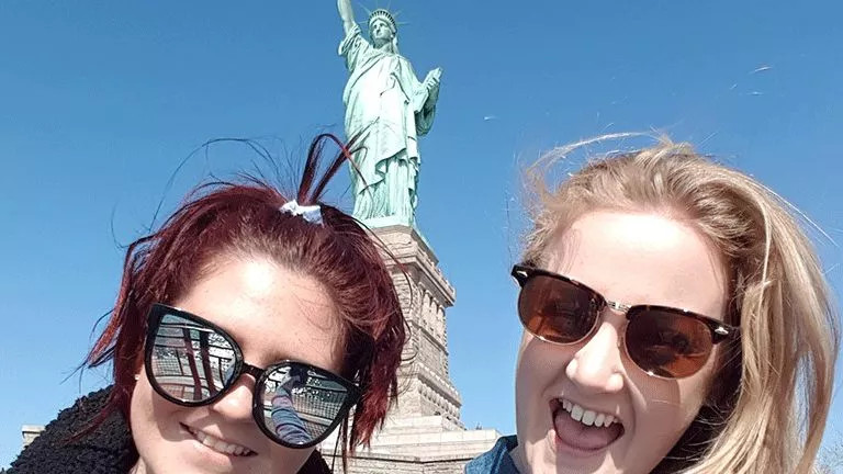 Two people take a selfie in front of the statue of Liberty.